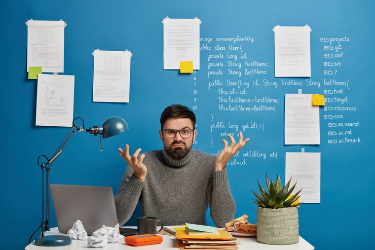 Bezpłatny plik Zdjęcie stresujący, profesjonalny maniak skupiony na monitorze nowoczesnego laptopa, nosi okulary optyczne, pozuje w przestrzeni coworkingowej na niebieskim tle