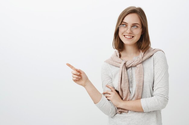 Strzał w górę kreatywnej, atrakcyjnej brunetki współpracowniczki w okularach i swetrze zawiązanym na szyi, patrząc i wskazując w lewo