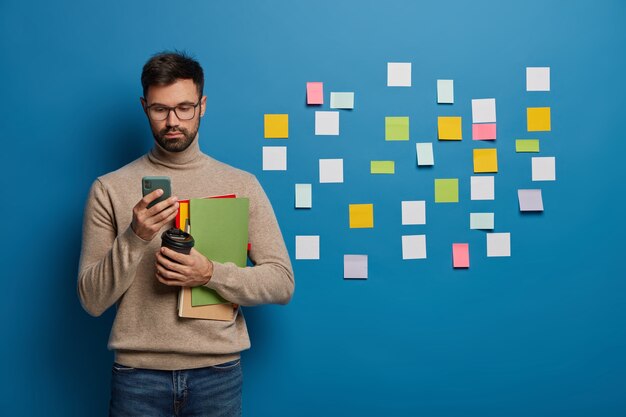 Student college'u używa telefonu komórkowego do rozmów online, pije kawę na wynos, trzyma notatniki lub podręczniki, przygotowuje się do lekcji, stoi za niebieską ścianą z wieloma samoprzylepnymi karteczkami