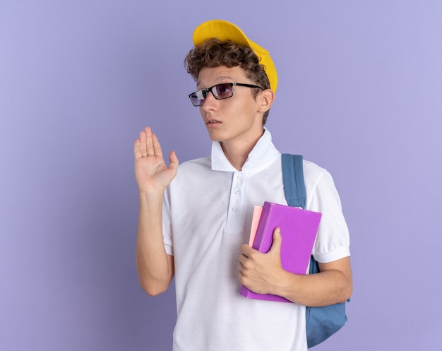 Student facet w białej koszulce polo i żółtej czapce w okularach z plecakiem trzymającym zeszyty, patrząc na bok zdezorientowany machający ręką stojącą na niebieskim tle