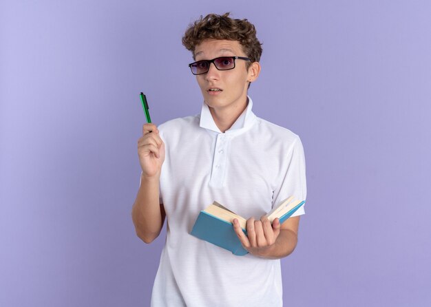Student facet w białej koszulce polo w okularach, trzymający książkę i długopis, patrzący w kamerę, zaskoczony, że ma nowy pomysł stojący na niebieskim tle