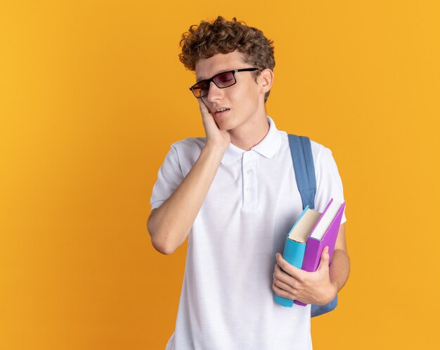 Student facet w zwykłym ubraniu, w okularach z plecakiem, trzymający książki, wyglądający na zmęczonego i znudzonego, z ręką na twarzy stojącej na pomarańczowym tle