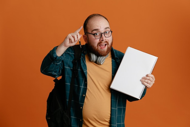 Student mężczyzna w zwykłych ubraniach w okularach ze słuchawkami i plecakiem, trzymający notatnik i długopis, patrzący na aparat, szczęśliwy i zaskoczony pokazujący palec wskazujący, mający świetny pomysł