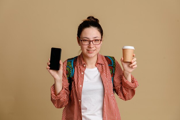 Student młoda kobieta w zwykłych ubraniach w okularach z plecakiem trzymająca filiżankę kawy i telefon komórkowy patrząc na kamerę szczęśliwy i pozytywny uśmiechnięty pewny siebie stojący na brązowym tle
