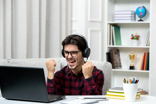 Student online młody facet w kraciastej koszuli i okularach studiujący na komputerze podekscytowany z pięściami w górę