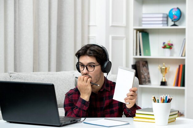 Student online słodki facet w kraciastej koszuli i okularach studiujący na komputerze gryzie palec