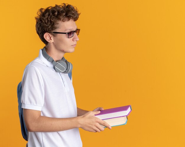 Student w swobodnym ubraniu ze słuchawkami w okularach i plecaku trzymającym książki holding