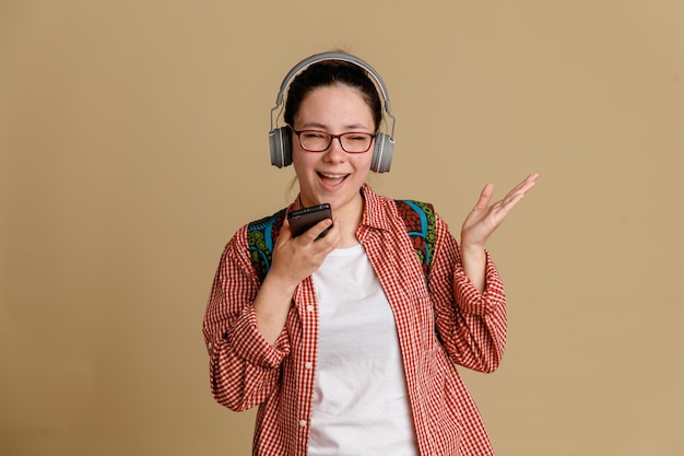 Studentka młoda kobieta w zwykłych ubraniach w okularach z plecakiem i słuchawkami na głowie trzymająca telefon komórkowy nagrywający wiadomość głosową szczęśliwy i pozytywny stojący na brązowym tle