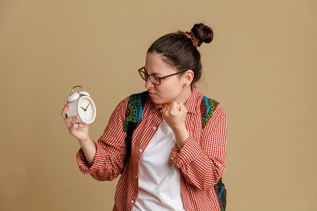 Studentka młoda kobieta w zwykłych ubraniach, w okularach z plecakiem, trzymająca budzik, patrząca na niego z gniewną twarzą, zaciskającą pięść w gniewie, stojącą nad brązowym tłem