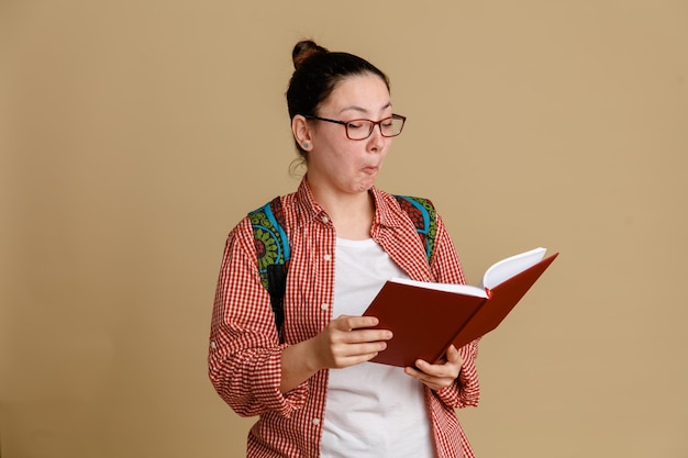 Studentka młoda kobieta w zwykłych ubraniach w okularach z plecakiem trzymająca notatnik patrząca zaintrygowana czytająca coś stojącego na brązowym tle