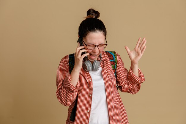 Studentka młoda kobieta w zwykłych ubraniach w okularach ze słuchawkami i plecakiem rozmawia przez telefon komórkowy, jest zła i szalona, stojąc na brązowym tle