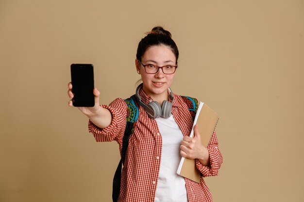Studentka młoda kobieta w zwykłych ubraniach, w okularach ze słuchawkami i plecakiem, trzymająca notebooka i telefon komórkowy pokazująca przed kamerą uśmiechający się pewnie stojący na brązowym tle