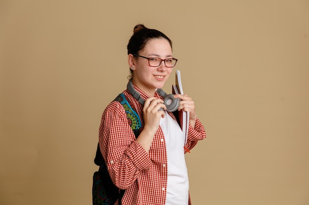 Studentka młoda kobieta w zwykłych ubraniach w okularach ze słuchawkami i plecakiem, trzymająca zeszyty, patrząca na kamerę, szczęśliwa i pozytywnie uśmiechnięta, pewna siebie, stojąca na brązowym tle