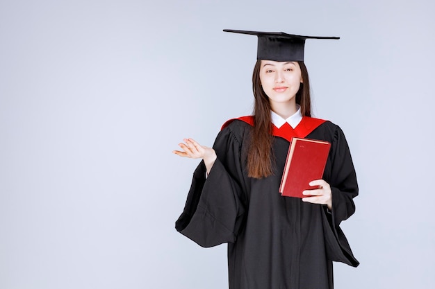 Studentka w akademickiej sukni trzymając czerwoną księgę i patrząc. zdjęcie wysokiej jakości