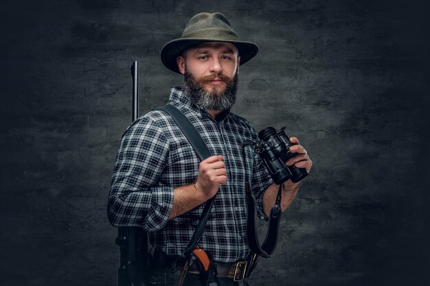 Studio portret brodatego mężczyzny myśliwego ubrany w koszulę z polaru w kratę posiada karabin.