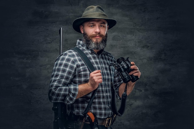 Studio portret brodatego mężczyzny myśliwego ubrany w koszulę z polaru w kratę posiada karabin.