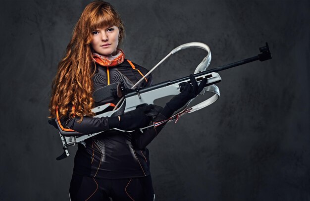Studio portret rudowłosej mistrzyni biathlonu trzyma broń na szarym tle.