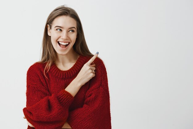 Studio portret uroczej zadowolonej młodej figlarnej kobiety w stylowym czerwonym luźnym swetrze, uśmiechając się szeroko, czując podniecenie, wskazując w prawym górnym rogu, stojąc