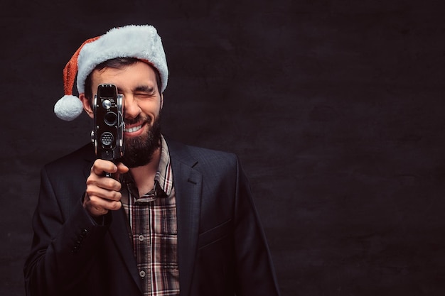 Studio portret uśmiechniętego brodatego mężczyzny w klasycznym garniturze i santa hat trzymając aparat retro, robiąc zdjęcie. Na białym tle na ciemnym tle.