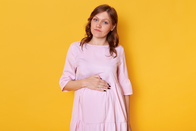 Studio strzelał młoda ładna kobieta w ciąży jest ubranym różową proszek suknię i trzyma rękę na brzuchu