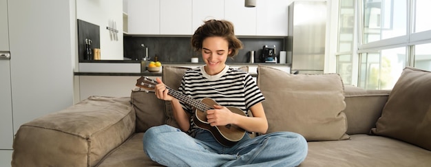 Bezpłatne zdjęcie styl życia i hobby koncepcja młoda uśmiechnięta kobieta na kanapie grająca na ukulele śpiewająca i ucząca się