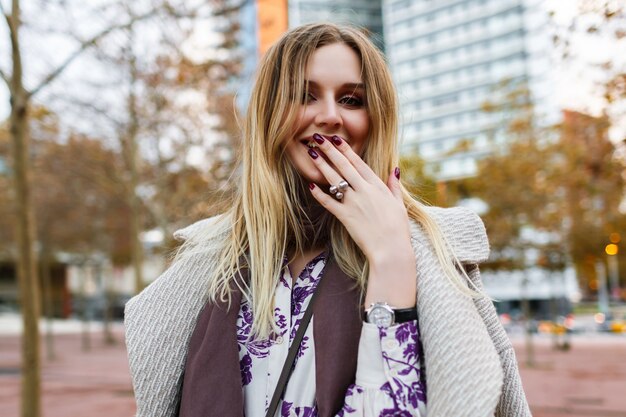 Styl życia na świeżym powietrzu pozytywny portret ładnej blondynki uśmiechniętej kobiety, która kończy się cieszyć czasem, elegancki strój glamour, ubrana w sukienkę i płaszcz, ciepłe kolory, wiosna jesień.