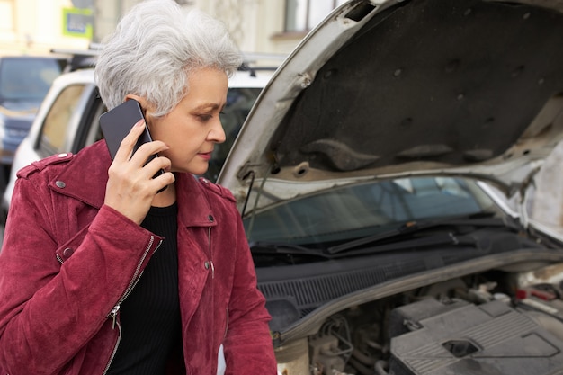 Bezpłatne zdjęcie stylowa atrakcyjna siwowłosa dojrzała kobieta kierowca stojąca obok zepsutego białego samochodu z otwartą maską i rozmawiająca przez telefon