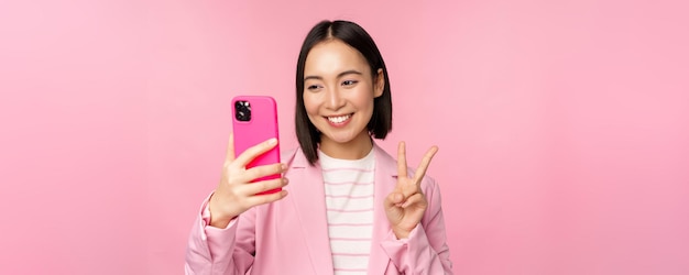 Stylowa azjatycka bizneswoman dziewczyna w garniturze biorąca selfie na czacie wideo na smartfonie z aplikacją telefonu komórkowego pozowanie na różowym tle studia