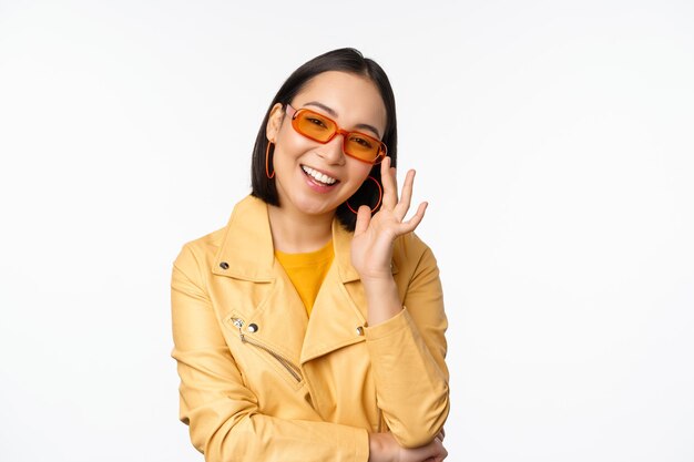 Stylowa azjatycka dziewczyna w okularach przeciwsłonecznych, uśmiechnięta i wyglądająca fajnie, stojąc na białym tle