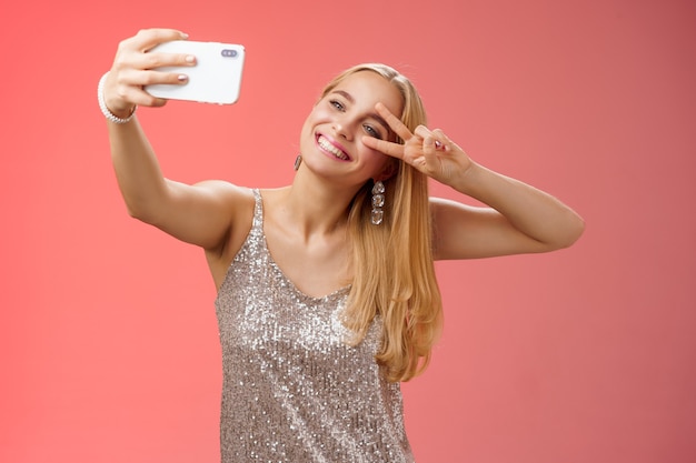 Stylowa, Bajeczna Glamour Młoda Blond Kobieta W Błyszczącej Srebrnej Sukience Przechylającej Głowę Beztroski Pokaz Pokoju Gest Znak Zwycięstwa Wydłużenie Ramienia Trzymającego Smartfona Przy Selfie Nagrywanie Wideo Post Online
