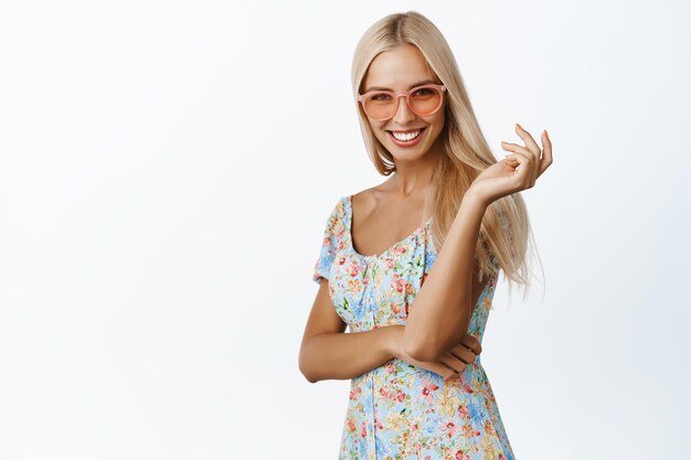 Stylowa blond letnia dziewczyna w okularach przeciwsłonecznych i sukience, uśmiechnięta i zalotna do kamery stojącej na białym tle