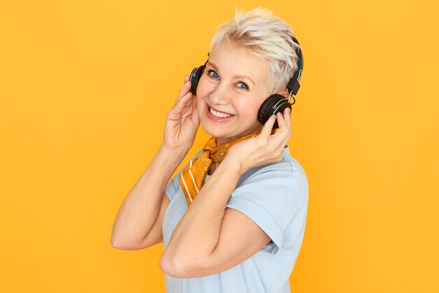 Stylowa blondynka na emeryturze ciesząca się muzyką, słuchająca radia w bezprzewodowych słuchawkach pozująca na żółto