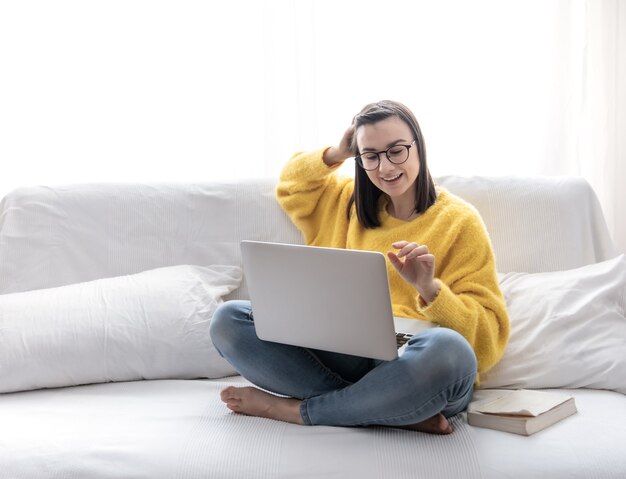 Stylowa brunetka w żółtym swetrze siedzi w domu na sofie w jasnym pokoju i pracuje zdalnie przy laptopie.