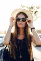 Bezpłatne zdjęcie stylowa dziewczyna stojąca w pobliżu drogi na sobie krótką czarną sukienkę, słomkowy kapelusz, czarne okulary i czarny plecak. uśmiecha się w ciepłych promieniach zachodzącego słońca. dotyka ręką okularów przeciwsłonecznych