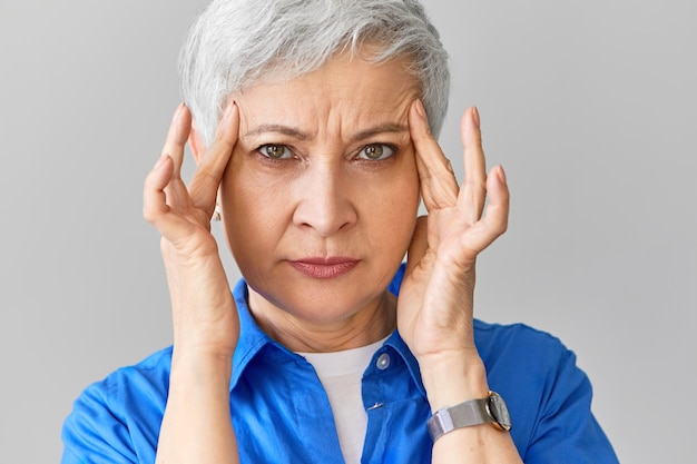Stylowa kaukaska kobieta w średnim wieku w niebieskiej koszuli cierpi na migrenę. Zbliżenie zestresowanej dojrzałej kobiety ściskającej skronie z powodu strasznego bólu głowy, masującej bolesne punkty