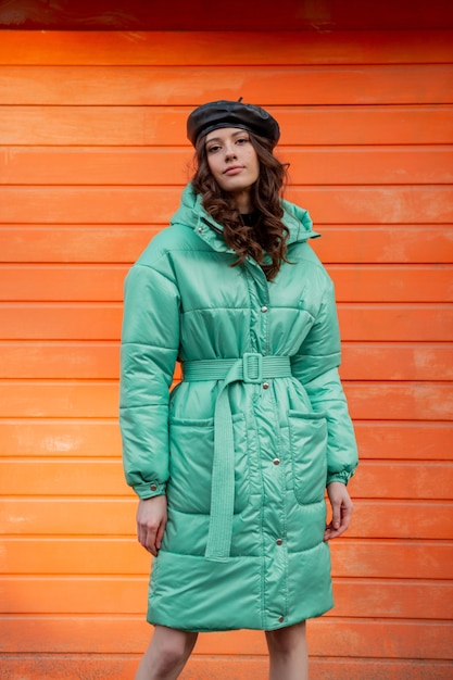 Bezpłatne zdjęcie stylowa kobieta pozuje zimą jesień trend w modzie puchowy płaszcz i kapelusz beret na pomarańczowej ścianie na ulicy