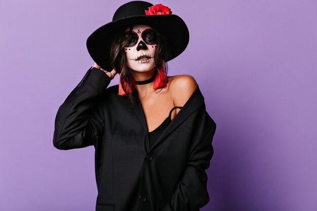 Stylowa kobieta w oversize'owej kurtce i nietuzinkowej halloweenowej masce dotyka ronda kapelusza. Portret atrakcyjna opalona dziewczyna na liliowej ścianie.