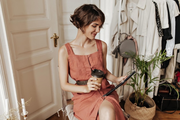 Stylowa krótkowłosa kobieta w lnianej sukience z czarnym paskiem uśmiecha się na krześle w przytulnym pokoju trzyma filiżankę kawy i tablet komputerowy