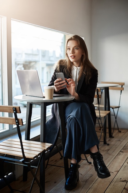 stylowa, ładna, nowoczesna kobieta w lokalnej kawiarni, siedząca przy oknie, pijąca kawę podczas pracy na laptopie, trzymająca smartfona, żeby zadzwonić do szefa