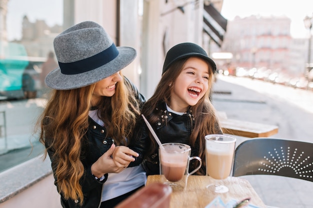 Stylowa mama i całkiem uśmiechnięta córka spędzają razem weekend w restauracji na świeżym powietrzu, pijąc kawę i koktajle mleczne.