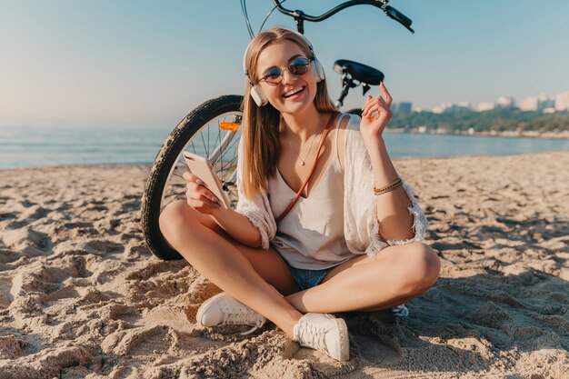 Stylowa młoda atrakcyjna blond uśmiechnięta kobieta siedzi na plaży z rowerem w słuchawkach, słuchając muzyki