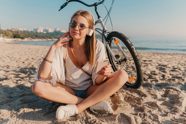 Stylowa młoda atrakcyjna blond uśmiechnięta kobieta siedzi na plaży z rowerem w słuchawkach, słuchając muzyki