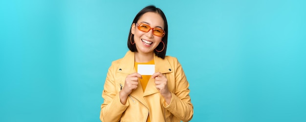 Bezpłatne zdjęcie stylowa młoda azjatka w okularach przeciwsłonecznych, pokazująca kartę kredytową i uśmiechnięta, polecająca bankowe płatności zbliżeniowe lub zniżki w sklepie, stojąca na niebieskim tle
