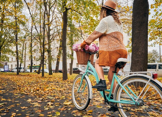 Stylowa młoda kobieta jedzie na rowerze z kwiatami na jesiennej ulicy