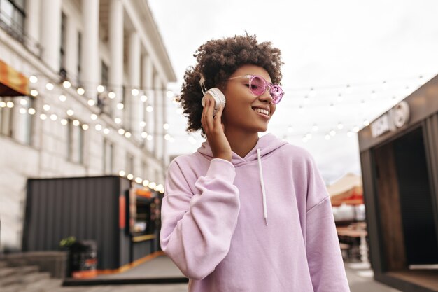 Stylowa młoda kręcona brunetka w różowych okularach przeciwsłonecznych i fioletowej bluzie z kapturem lubi muzykę w słuchawkach i uśmiecha się na zewnątrz