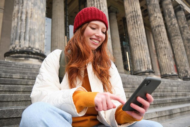 Stylowa młoda ruda kobieta rozmawiająca przez aplikację na telefon komórkowy za pomocą aplikacji społecznościowej szuka s