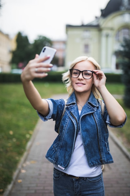 Stylowa moda blondynka kobieta w dżinsach i okularach sprawia, że selfie na swoim telefonie w mieście rano