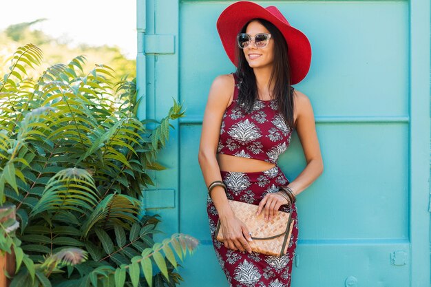 Stylowa piękna kobieta w czerwonym kapeluszu pozująca na niebieskiej ścianie, drukowany strój, letni styl, trend w modzie, top, spódnica, chuda, słomkowa torebka, okulary przeciwsłoneczne, akcesoria, uśmiechnięta, szczęśliwa, tropikalne wakacje