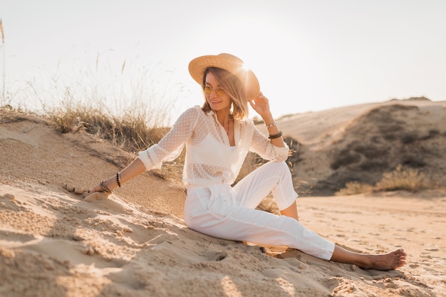 Stylowa piękna kobieta w piasku plaży pustyni w białym stroju na sobie słomkowy kapelusz na zachód słońca