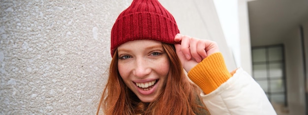 Bezpłatne zdjęcie stylowa ruda dziewczyna w czerwonym kapeluszu uśmiecha się i wygląda na szczęśliwą, pozując na zewnątrz na ulicy, wygląda na zrelaksowaną i